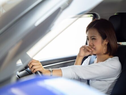 a lady in car have a hand on her nose, seems like she is smelling gasoline smell in her car.